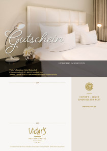 Ein elegantes weißes Bettkopfteil ist zu sehen mit doppelten Kissen. Im Vordergrund ein Nachttisch mit Nachttischlampe und Blumendekoration.